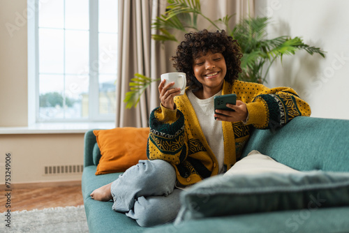 Good morning. African girl drinking coffee holding smartphone sitting on couch at home Woman with cell phone surfing internet using social media apps. Shopping online Internet news cellphone addiction
