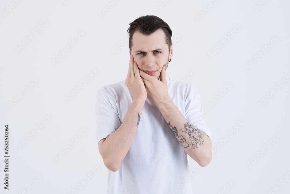 Attractive Caucasian young man with tattoos in casual clothes touching cheek and suffering due to toothache isolated on white studio background. Attractive Caucasian young man with tattoos in casual clothes touching cheek and suffering due to toothache isolated on white studio background.
