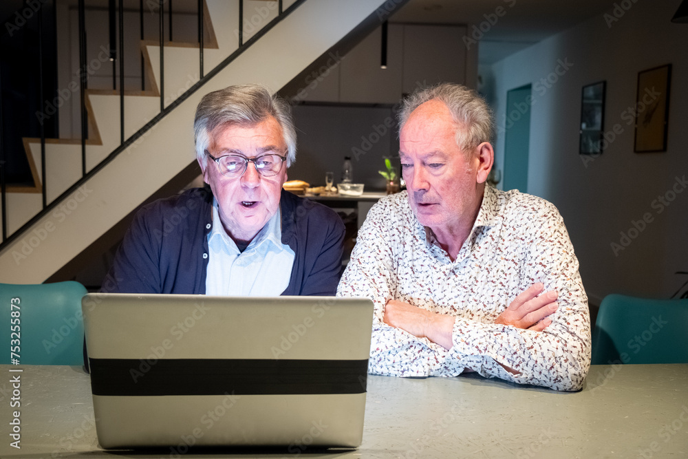 In this image, two senior men are engrossed in viewing a laptop screen. The man on the left, with his glasses slightly down his nose and a dark jacket over a blue shirt, exhibits an expression of In this image, two senior men are engrossed in viewing a laptop screen. The man on the left, with his glasses slightly down his nose and a dark jacket over a blue shirt, exhibits an expression of