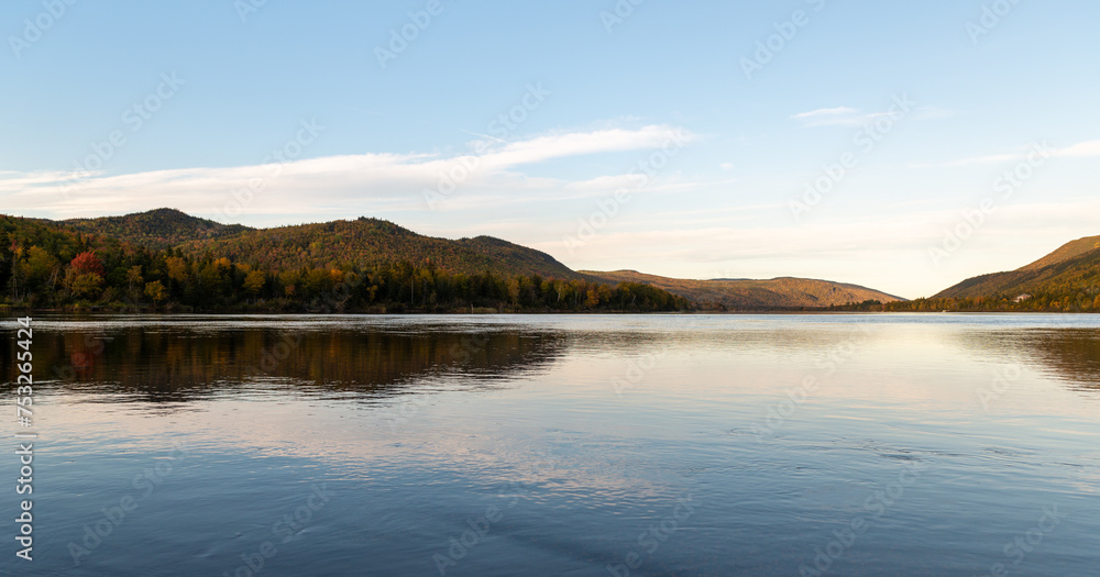 A calm and scenic view of mountains covered in red and orange colored trees. The fall colors are reflected in the smooth blue ocean. The sky has an orange hue as it sets behind the hills. 