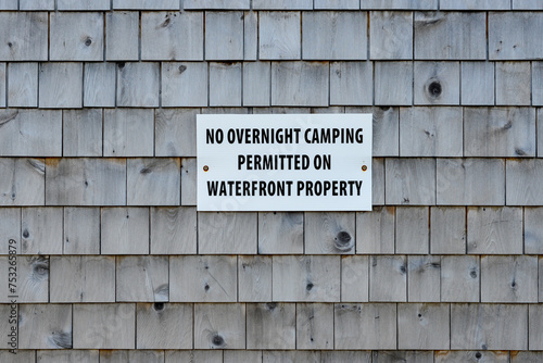 No overnight camping permitted on waterfront property sign posted on a grey cedar shake exterior wall. The message restricts access to shoreline property. The text is black on a small white board.