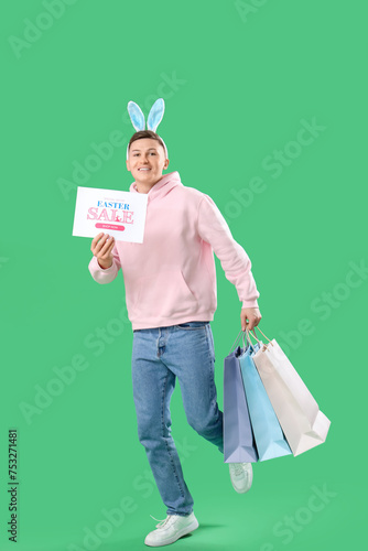 Running young man in bunny ears holding paper with text EASTER SALE and shopping bags on green background