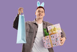 Young man in bunny ears with gifts and shopping bag on lilac background. Easter Sale