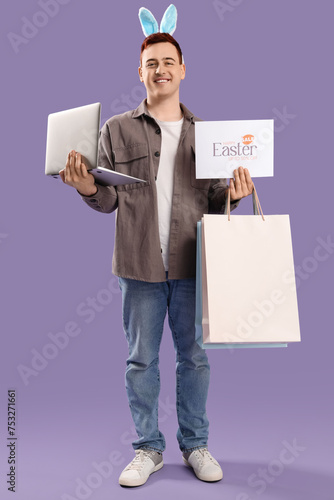 Young man in bunny ears with laptop and shopping bags on lilac background. Easter Sale