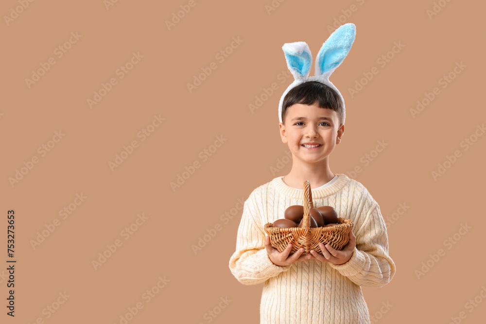 Fototapeta premium Cute little boy in bunny ears headband holding basket with chocolate eggs for Easter on beige background