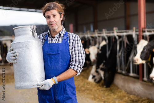 Skilled middle-aged male farmer carrying milk churn near stall with cows during work on dairy farm © JackF