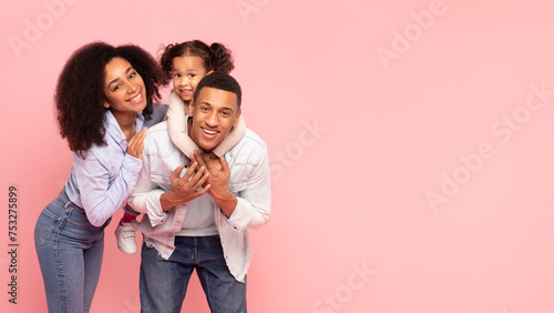 Happy african american family of three, man giving piggy back ride for his daughter, smiling together at camera over pink background, panorama, free space