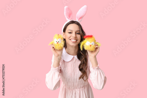 Happy young woman in Easter bunny ears headband with toy chickens on pink background