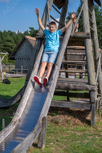 A boy in a blue t-shirt and denim shorts slides down a wooden playground slide, arms raised with exhilaration. Perfect for themes of joy, outdoor play, and childhood adventures.