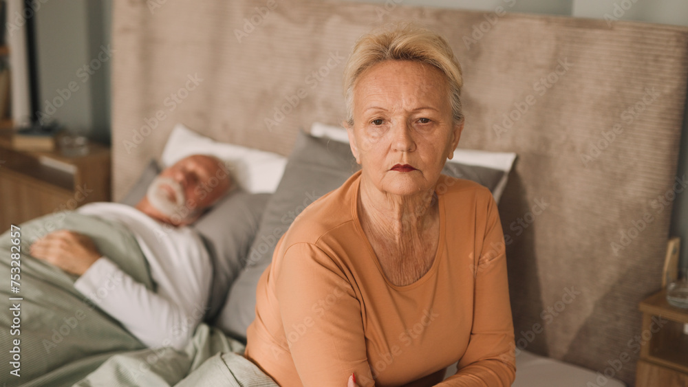 Worried senior woman sitting on bed with her husband sleeping in background Worried senior woman sitting on bed with her husband sleeping in background