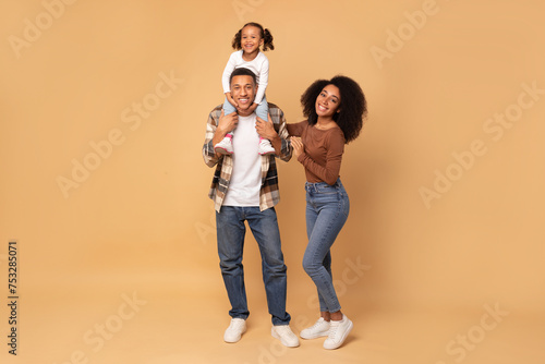 Happy black parents and little daughter sitting on dad's shoulders, having fun and posing together over peach studio background, full length shot © Home-stock