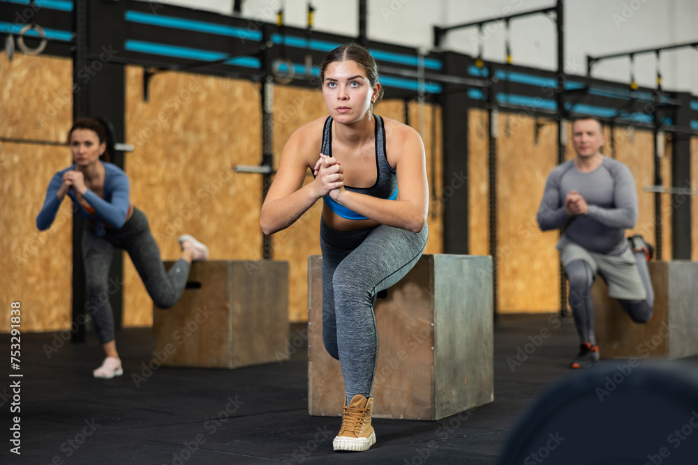 Concentrated young girl in fitness attire performing Bulgarian split squats using wooden plyometric box at gym. Bodyweight training concept.. - obrazy, fototapety, plakaty 