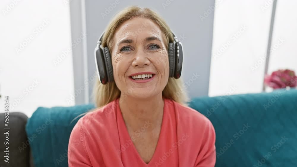 Smiling middle-aged blonde woman wearing headphones at home, exuding a friendly mature beauty in a comfortable indoor setting. Smiling middle-aged blonde woman wearing headphones at home, exuding a friendly mature beauty in a comfortable indoor setting.