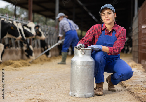 Female farmer holding large metallic milk can in hangar with cows