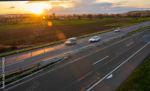 Cars drive at high speed on the highway through the rural landscape. Fast blurred highway driving. A scene of speeding on the highway. Beautiful sunset in the background. 