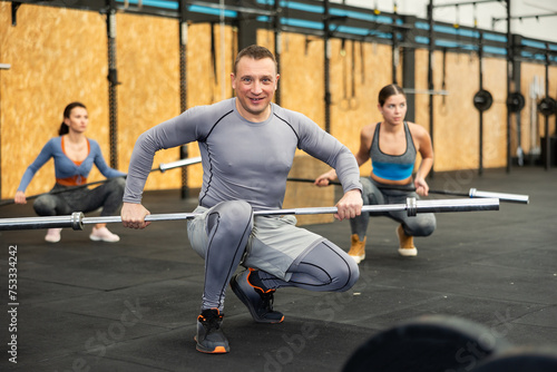 Smiling athletic man squatting with empty barbell bar in hands, preparing to perform exercises during intense group training at modern gym