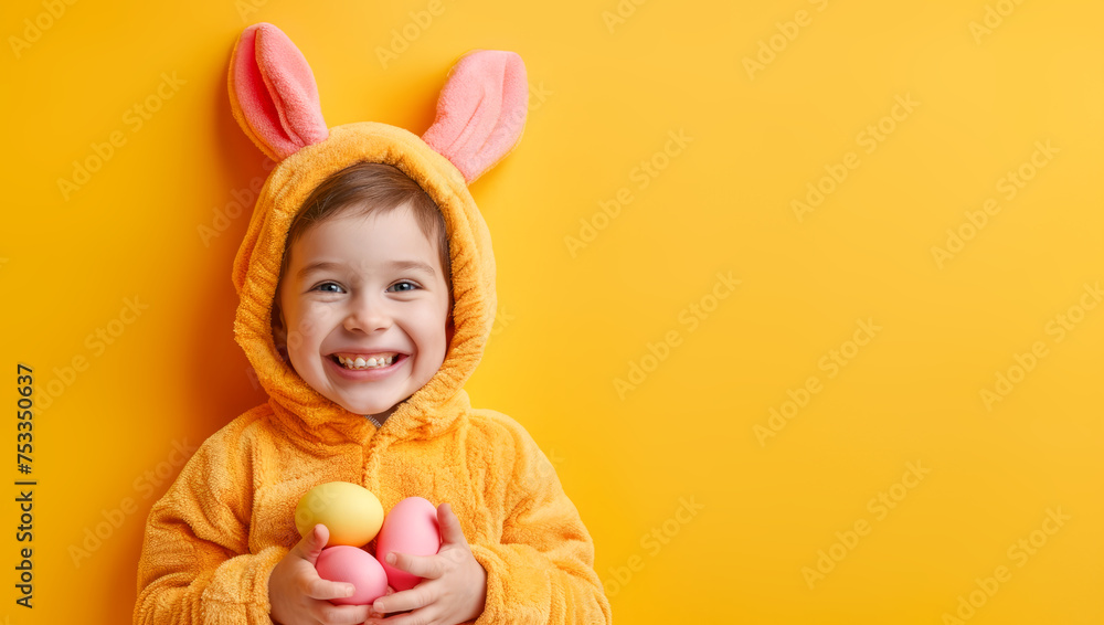 Funny happy child boy in bunny costume holding easter eggs in hands on yellow background. Smiling kid with rabbit ears.