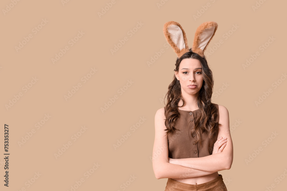 Sad young woman in Easter bunny ears headband on beige background