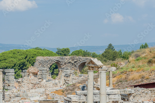 Ruins of an ancient city with a prominent archway and columns amid green landscapes, in Ephesus, Turkiye photo