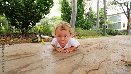 Cheeky toddler lies down on ground after runbike slip, dramatically pretending to be injured. With exaggerated and mischievous expression, he awaits sympathy for minor incident. Camera move closer.