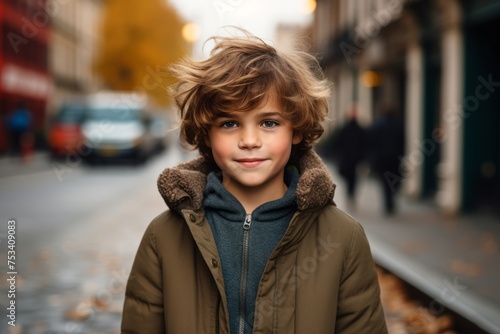 Outdoor portrait of cute little boy with blond curly hair, wearing warm clothes, posing on the street.