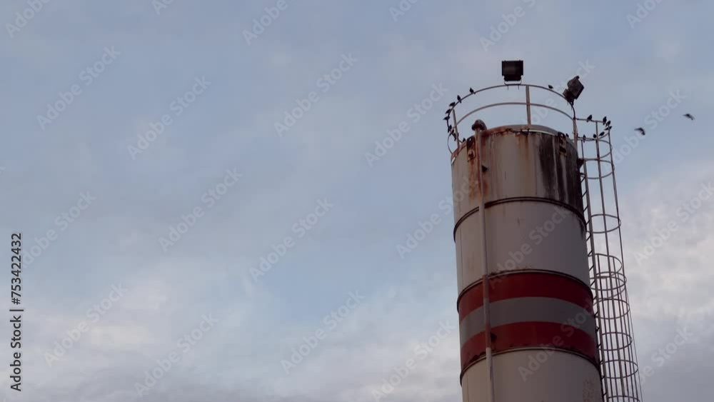 An industrial chimney stands tall against a cloudy sky, with birds perched atop it on wires. An industrial chimney stands tall against a cloudy sky, with birds perched atop it on wires.