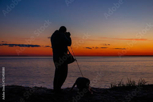 Silhouette of a guy with photo camera and dog on a river shore at sunset