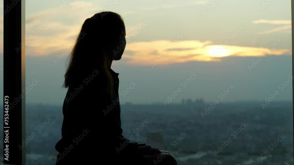 Woman sits by window in dimly lit room, gazing out at city in late evening. Atmosphere is introspective, capturing mood of uncertainty and solitude. Blurred cityscape seen below in twilight Woman sits by window in dimly lit room, gazing out at city in late evening. Atmosphere is introspective, capturing mood of uncertainty and solitude. Blurred cityscape seen below in twilight