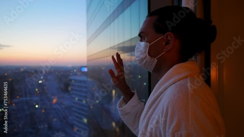 In dimly lit hotel room, man in face mask stands by window, hand on glass, gazing at evening cityscape during quarantine. Room is silent, and he is alone, thoughtful, contemplating city below photo