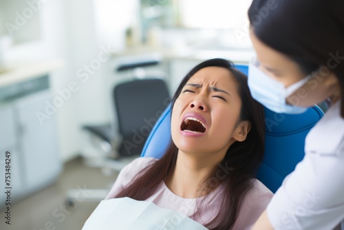 Portrait of a young woman in her late 20s, of Asian descent, grimacing in pain while seated in a dentist's chair, complaining about toothache, with the dentist intentionally blurred in the back
