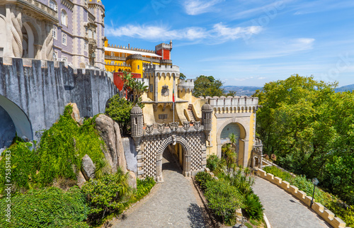 Aerial panoramic exterior and wide view of the colorful Pena Palace with the path up to the arched entrance decorated with bas-reliefs, under a sunny blue sky. Sintra. Portugal.
