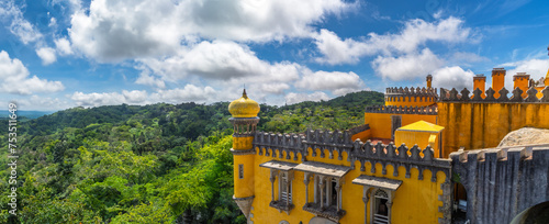 Wide angle exterior view of the top of the yellow watchtower and attic of the Pena Palace, under a sunny cloudy blue sky, with the leafy Sintra Mountains below on the horizon. Portugal.