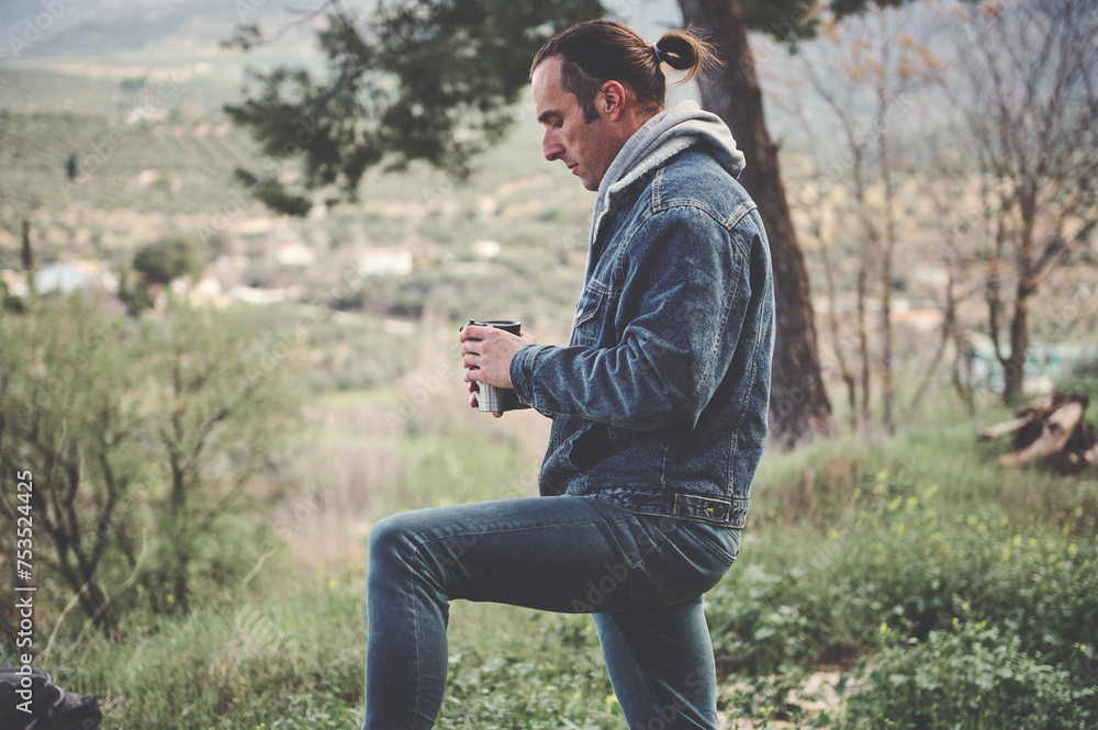 Handsome young man in casual denim clothes, standing in the forest alone with a thermos mug of hot drink in his hands Handsome young man in casual denim clothes, standing in the forest alone with a thermos mug of hot drink in his hands
