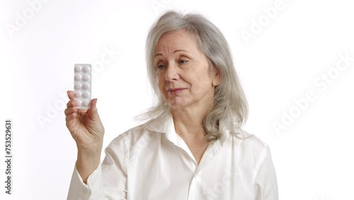 Elderly lady with gray hair, holding a blister pack of pills, showing doubt and skepticism. Wearing white blouse, isolated on white background. Perfect for medical adherence concepts. Camera 8K RAW. photo