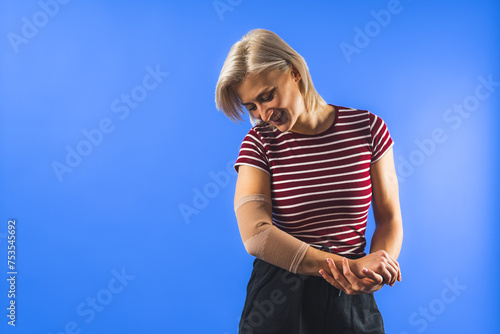 medium shot of a young woman looking at a bandage on her elbow for support, blue background copy space. High quality photo