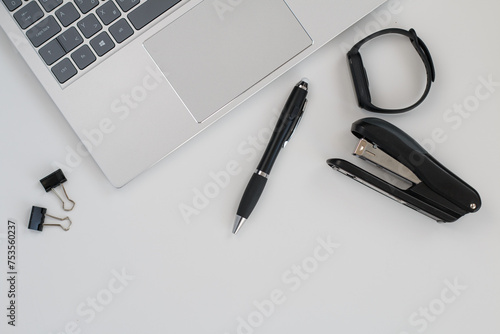 A silver laptop with a black pen, a black smartwatch and a stapler on an office desk, top view. 