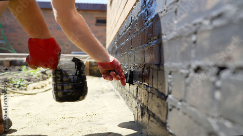 A worker applies waterproofing to the fa ade of a house with a brush. Close-up of a basement brush painted black. House renovation, wall painting, moisture protection.