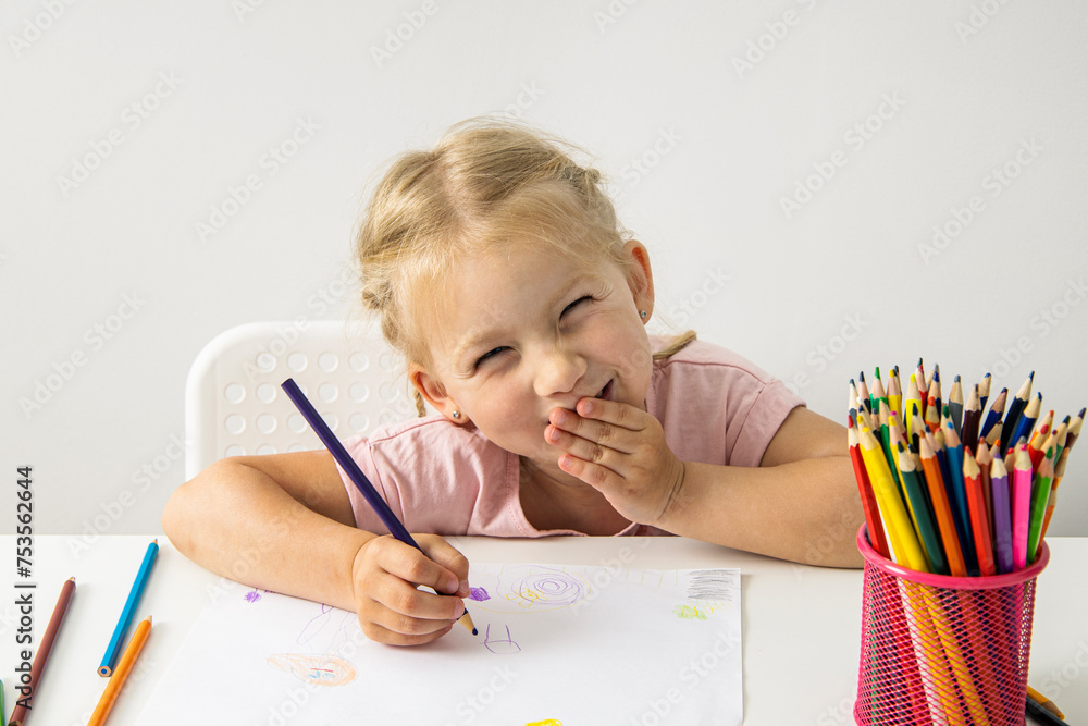 A smiling child blonde girl draws with colored pencils sitting at a white table A smiling child blonde girl draws with colored pencils sitting at a white table