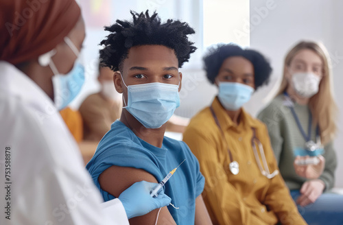 A nurse is giving support to a young man getting a shot in a clinic, with people wearing face masks in the background