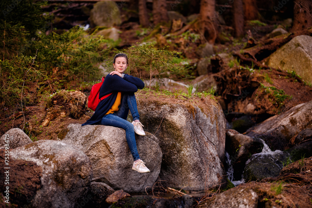 Obraz premium Amidst a rustic forest setting, a young woman finds a moment of reflection while seated on a moss-covered boulder
