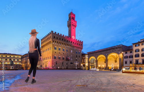 A beautiful blonde woman with a stylish hat wearing black tight trousers walking on the street - Palazzo Vecchio or Palazzo della Signoria at twilight blue hour - Florence, Italy.  photo