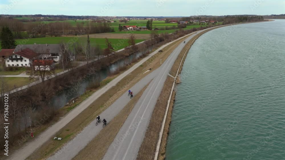 Cyclists ride on an embankment bicycle path along River Innnear Rosenheim, Bavaria, Germany. Am Innradweg bei Rosenheim. Gravel bike path along a wide river in Deutschland. Cycling in upper Bayern. Cyclists ride on an embankment bicycle path along River Innnear Rosenheim, Bavaria, Germany. Am Innradweg bei Rosenheim. Gravel bike path along a wide river in Deutschland. Cycling in upper Bayern.