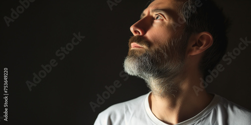 Pensive bearded man looking upwards, half face illuminated by dramatic lighting on a dark background