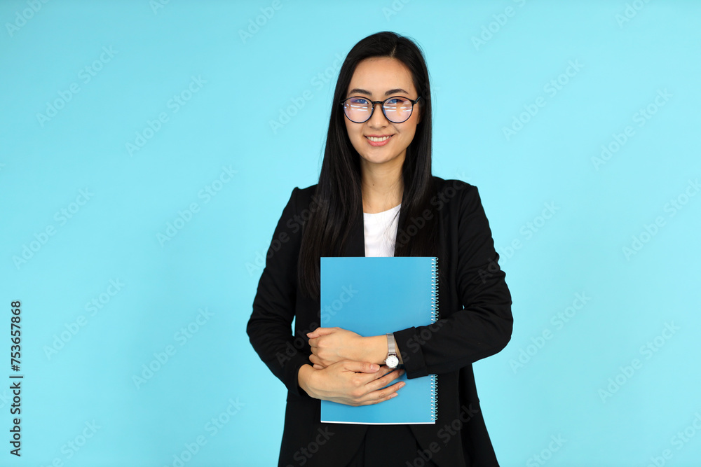 An Asian woman, against a blue background, holds documents, embodying confidence and academic success. An Asian woman, against a blue background, holds documents, embodying confidence and academic success.