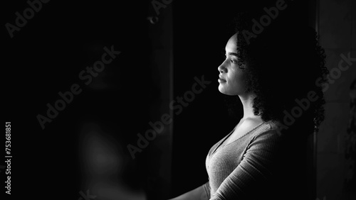 One thoughtful young black woman gazing out by window at home in monochromatic, black and white. Profile face of an African American person with pensive contemplative emotion
