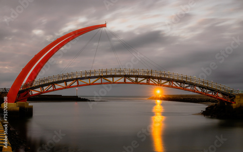 Fototapeta Naklejka Na Ścianę i Meble -  A gloomy sunset in a cloudy evening was sparkled by the light house beneath a red arch bridge