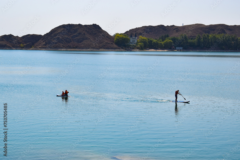 The bright summer sun illuminates the clear water of lake Kapchagay with a rocky shore, people are swimming on SUP boards, and in the distance you can see a house surrounded by dense trees. Kazakhstan