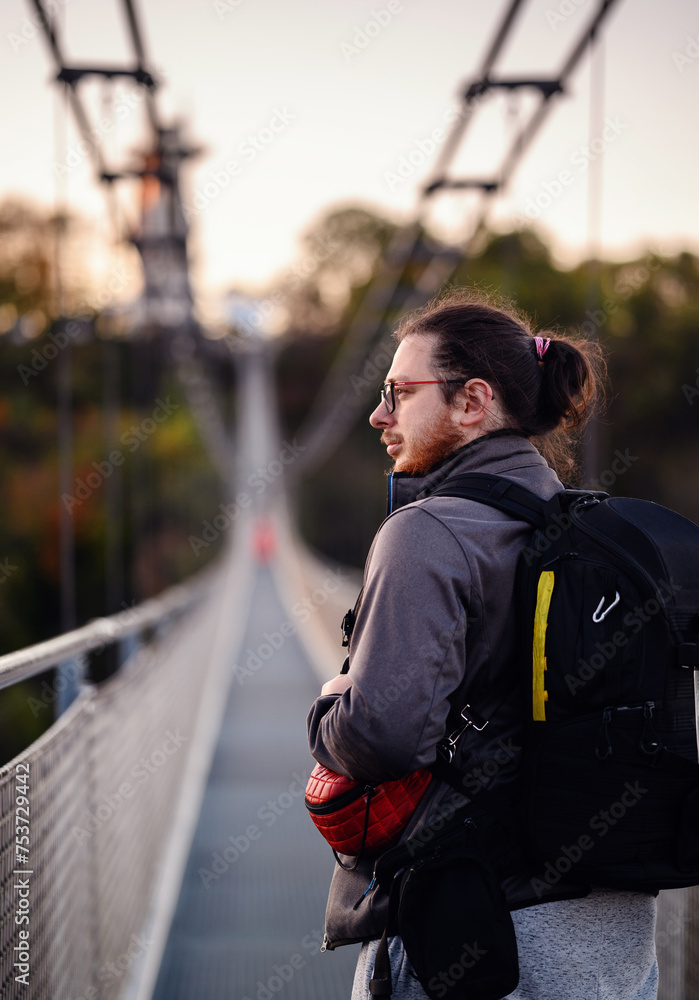 A pensive traveler with a backpack takes a moment to gaze into the distance on a suspension bridge, enveloped by the soft hues of dusk and autumn foliage