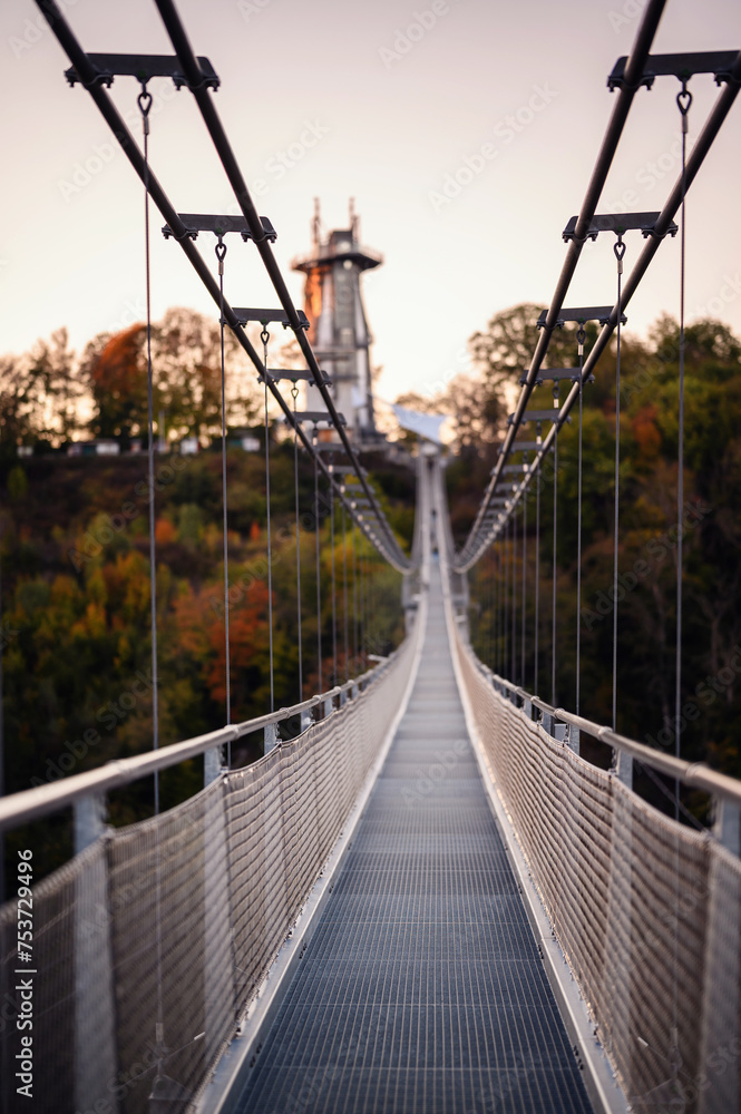 Fototapeta premium A suspension bridge stretches into the distance, framed by the warm light of dawn reflecting off the autumn-colored trees and the calm water below