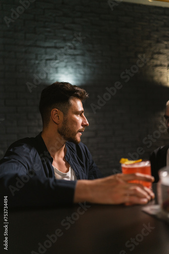 Young bearded man sits at bar counter holding alcoholic cocktail. Man enjoys evening party vibe while taking moment to relax